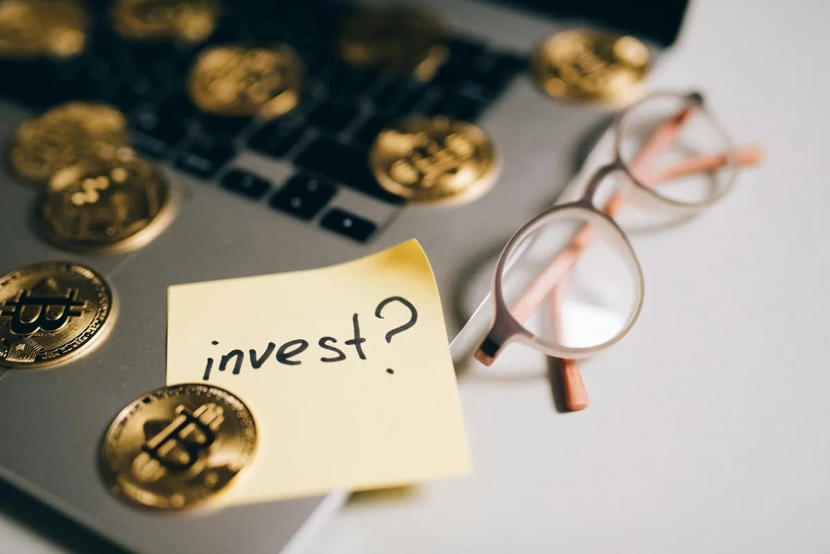 A close-up of bitcoin coins on a laptop with eyeglasses and a sticky note reading 'invest?'
