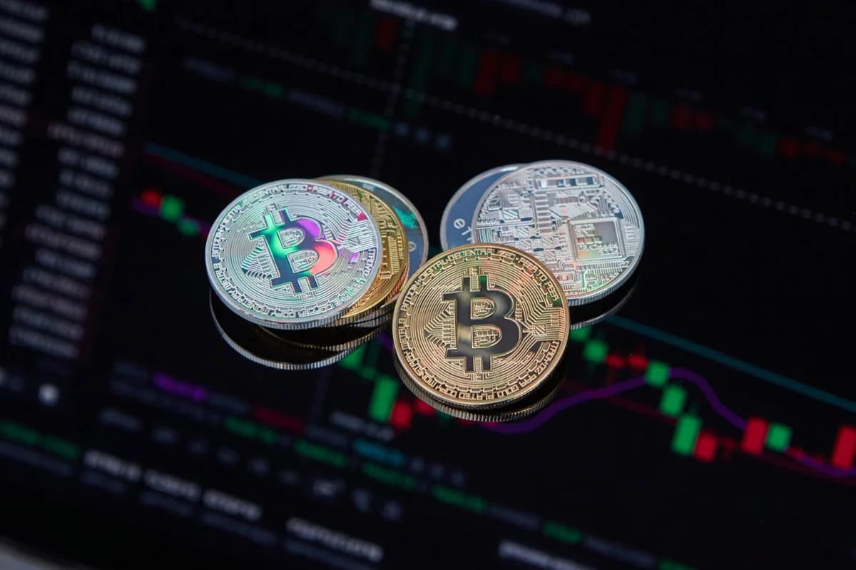 A hand holds an Ethereum coin in front of a laptop displaying cryptocurrency trading charts.