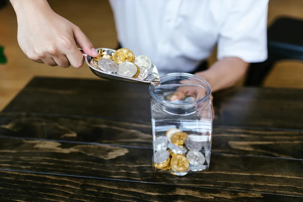 Close-up of hands scooping assorted cryptocurrency coins into a jar on a wooden surface.