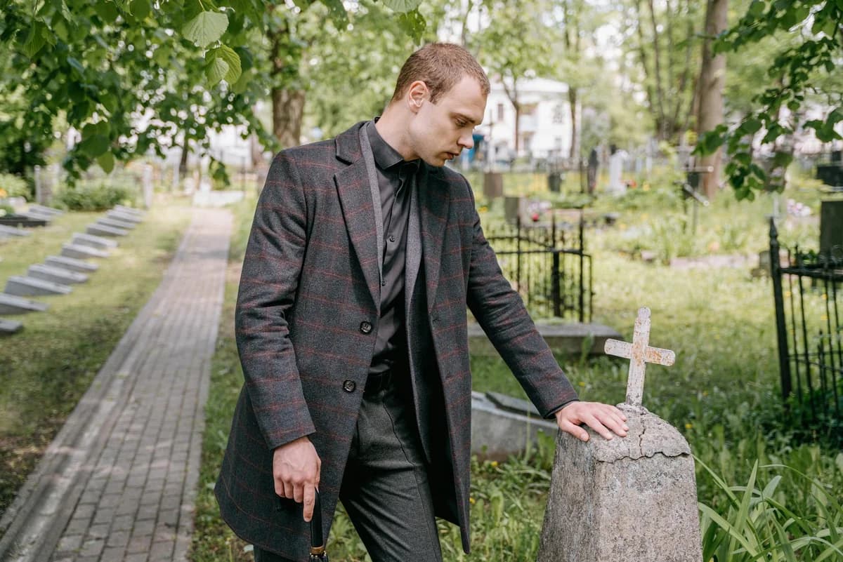 A man in a coat stands solemnly by a gravestone in a cemetery on a sunny day.