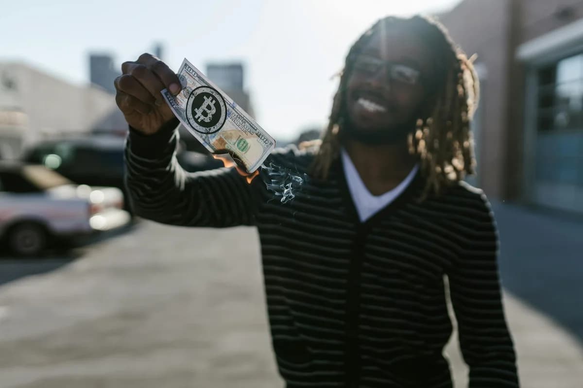 A man with dreadlocks holds a burning bill featuring a bitcoin logo, symbolizing cryptocurrency volatility.