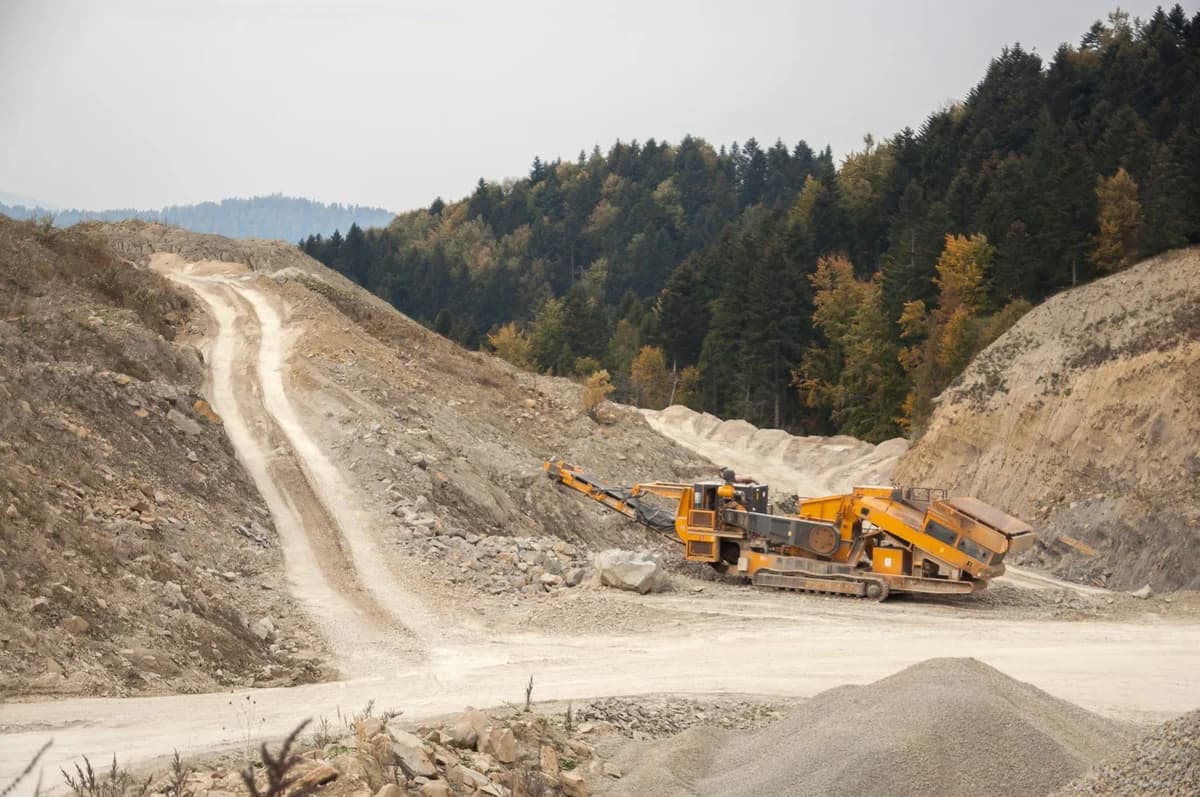 Industrial crusher machine reducing rocks near big pile of soil with tracks in nature near woods with lush green trees