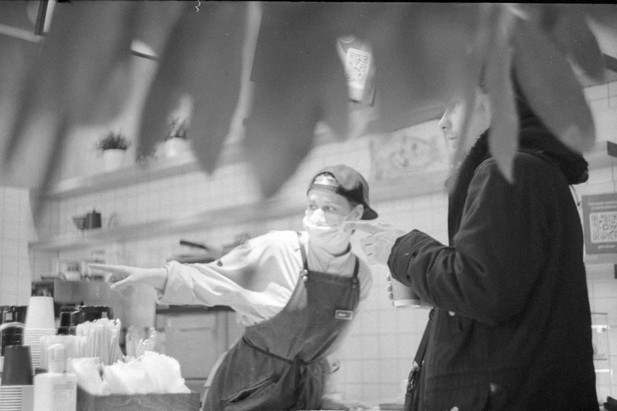 Masked café worker in apron assists a customer inside a modern setting. Safety focused.