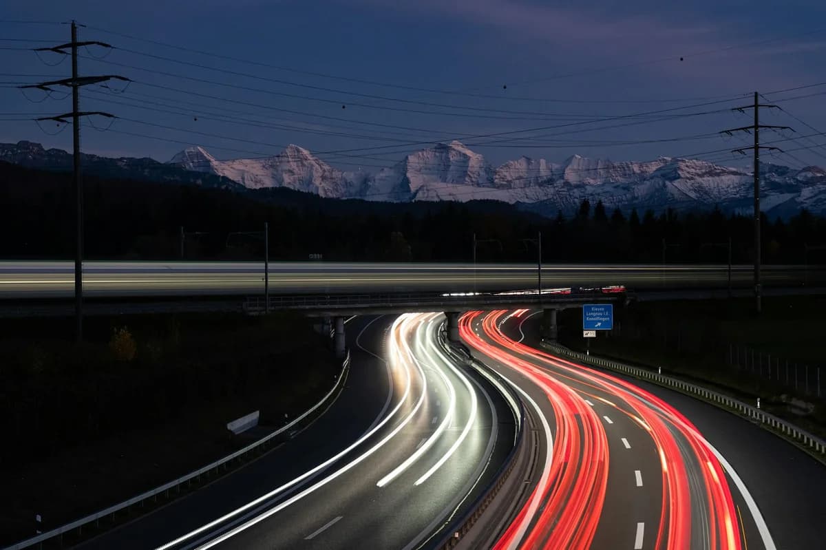 Night shot of a highway in Bern with moving car light trails and snowy mountain backdrop.