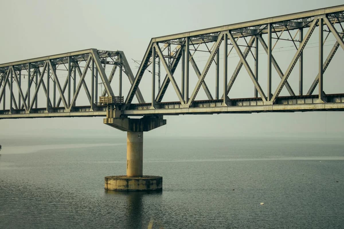 Metal net barrier near modern long suspension stone bridge with metal constructions crossing calm sea on city with bright sunshine