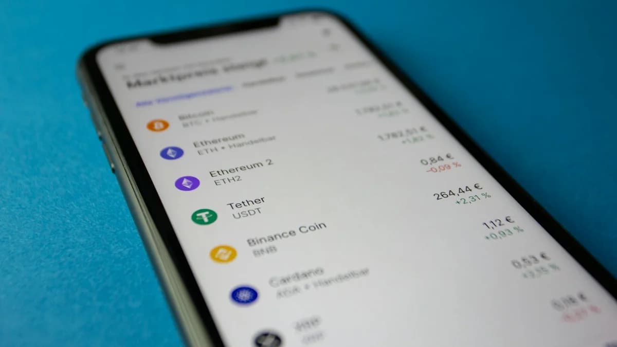 A person checks cryptocurrency values on a smartphone with a laptop and Bitcoins nearby.