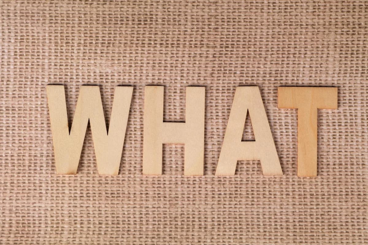 Wooden letters forming the word WHAT set on a textured burlap surface.