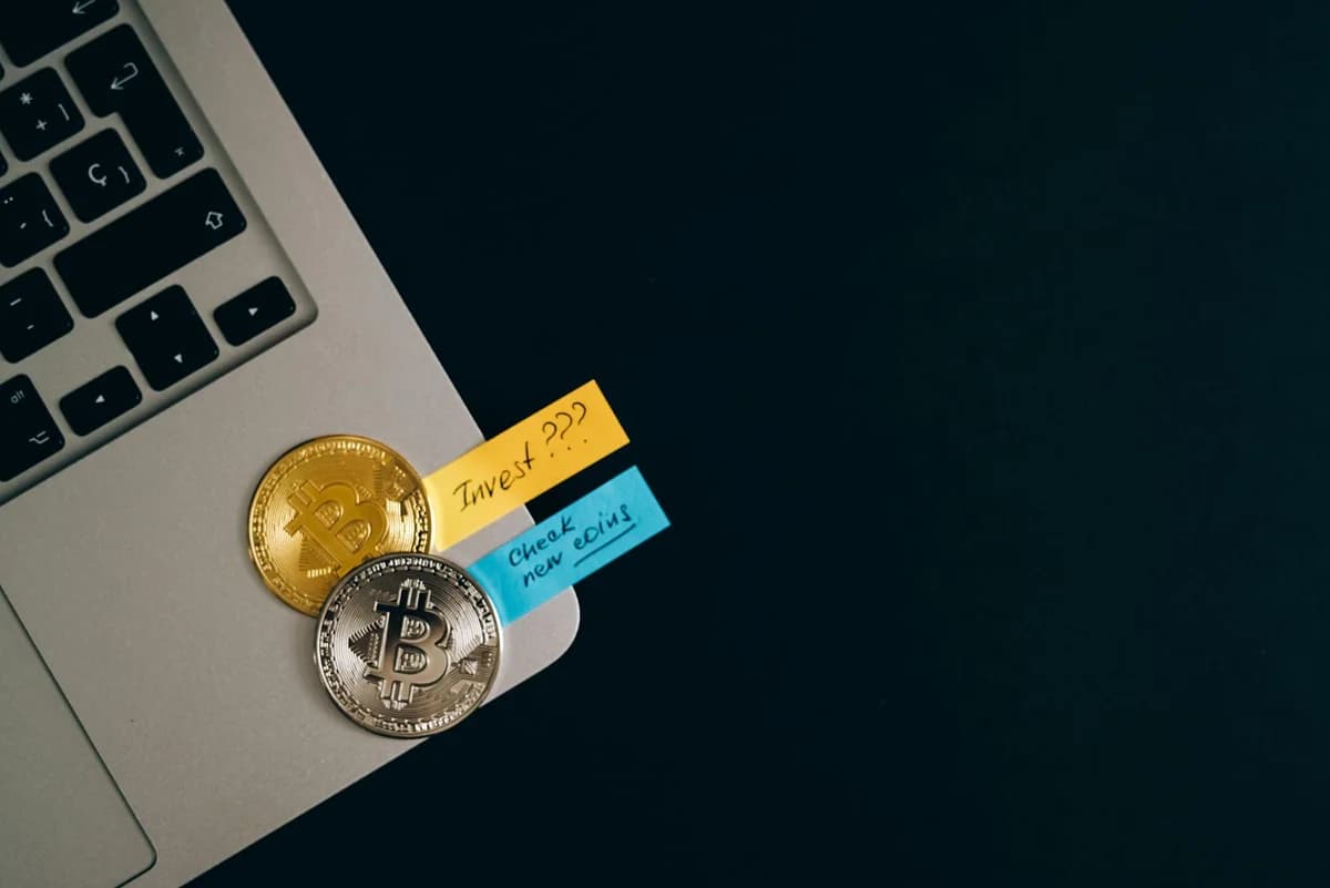 Bitcoin coins placed on a laptop keyboard with sticky note reminders about investing and checking new coins.