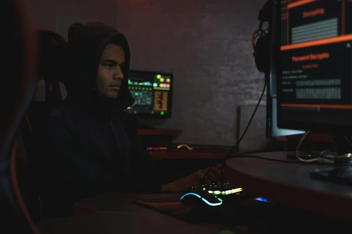 Masked individual in dark room interacting with computer screens displaying hacker imagery and code.