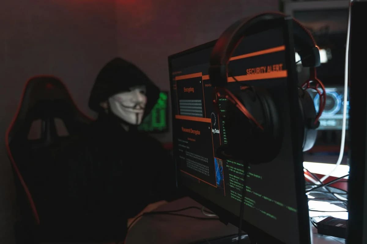 What Is the Biggest DeFi Hack of All Time? A person wearing a Guy Fawkes mask engaged in hacking activities on a computer in a dimly lit room.